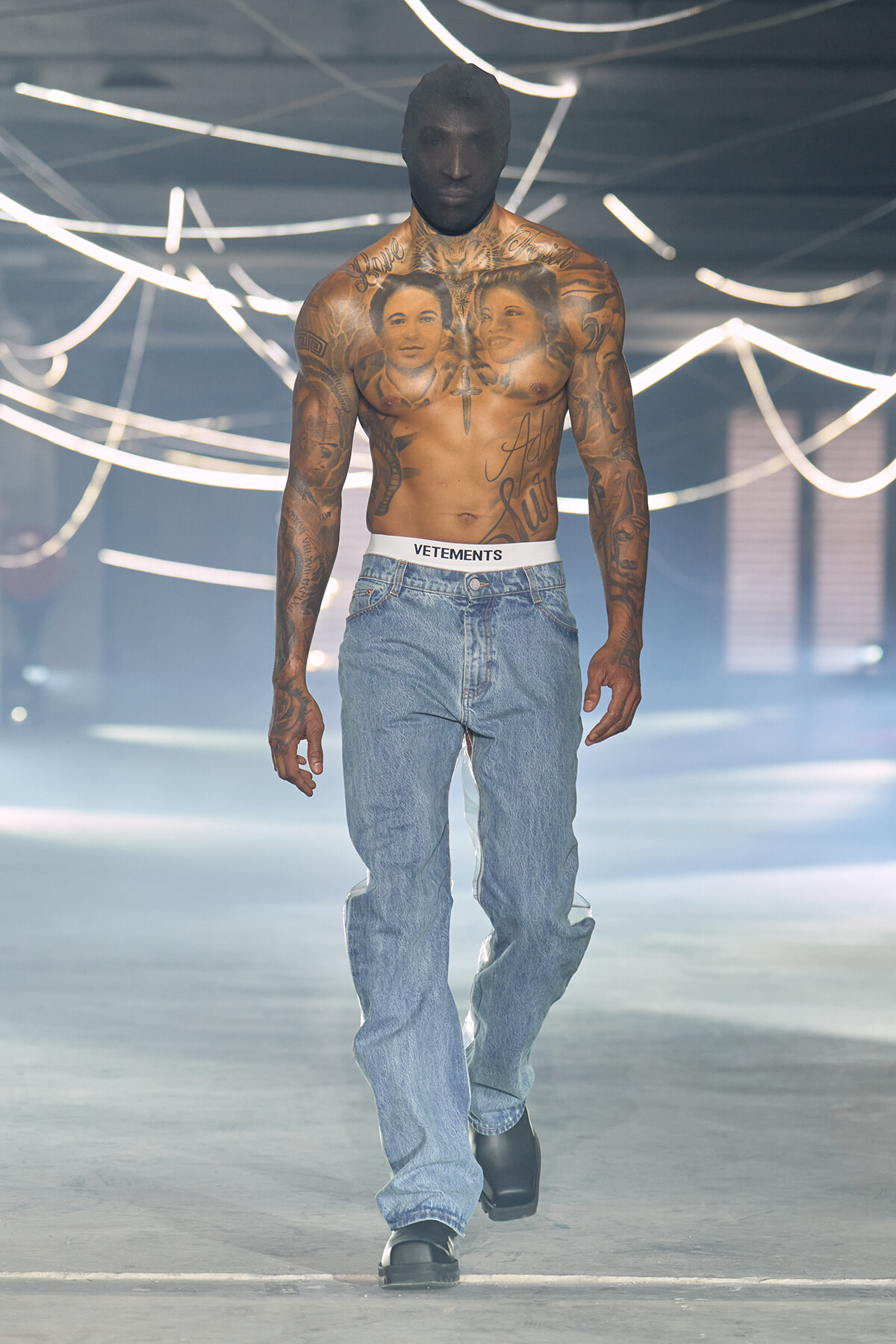Tall shirtless male model with extensive chest and arm tattoos walking a fashion runway in blue jeans and a white Vetements waistband; neon light backdrop.