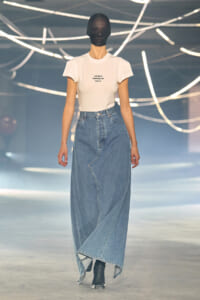 Model walking a runway in a white fitted T-shirt tucked into a high-waisted, asymmetrical denim maxi skirt with black ankle boots.