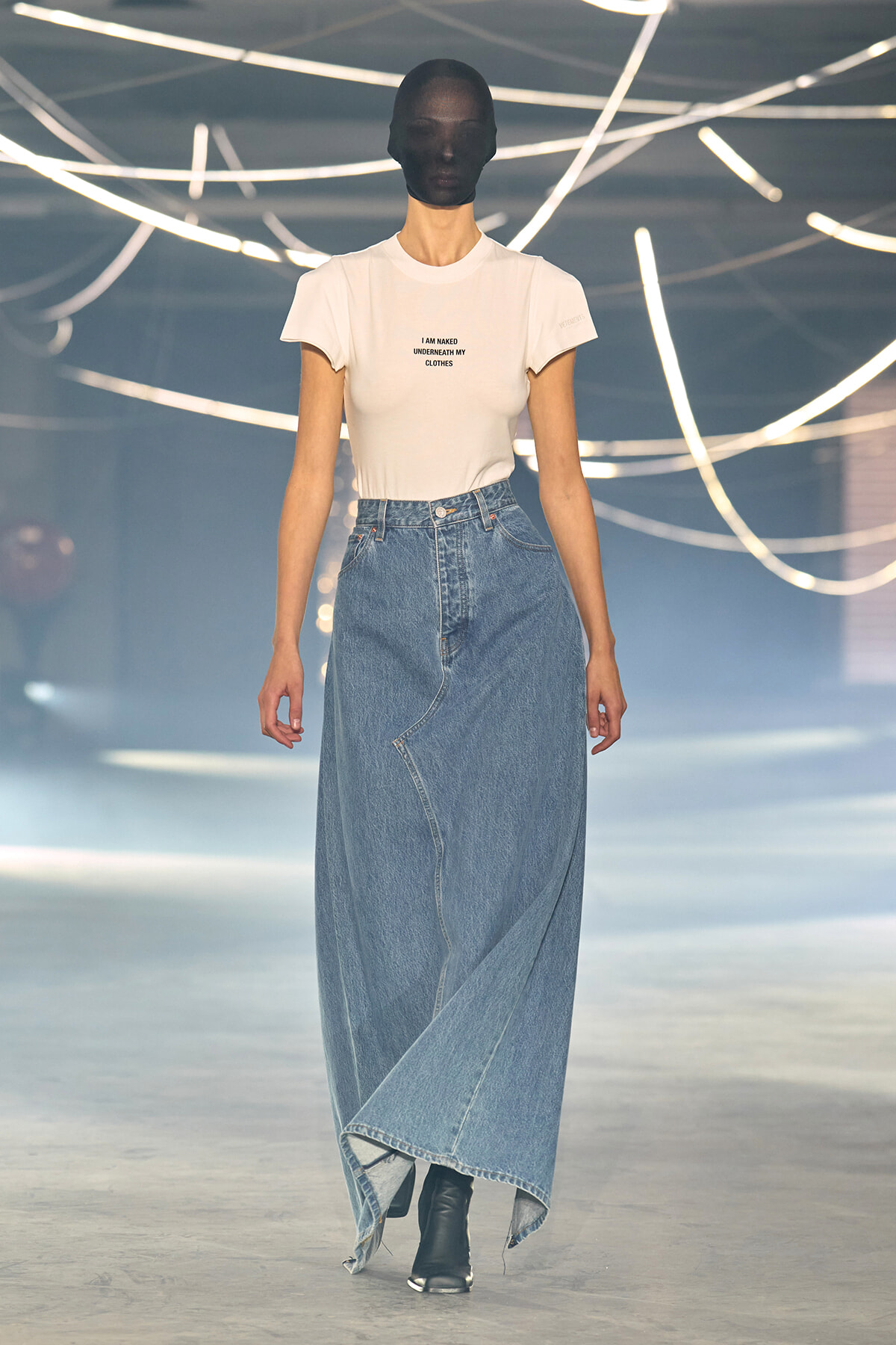 Model walking a runway in a white fitted T-shirt tucked into a high-waisted, asymmetrical denim maxi skirt with black ankle boots.