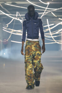 Male model walking the runway in a navy zip-up jacket and camouflage cargo pants, with neon light tubes overhead.