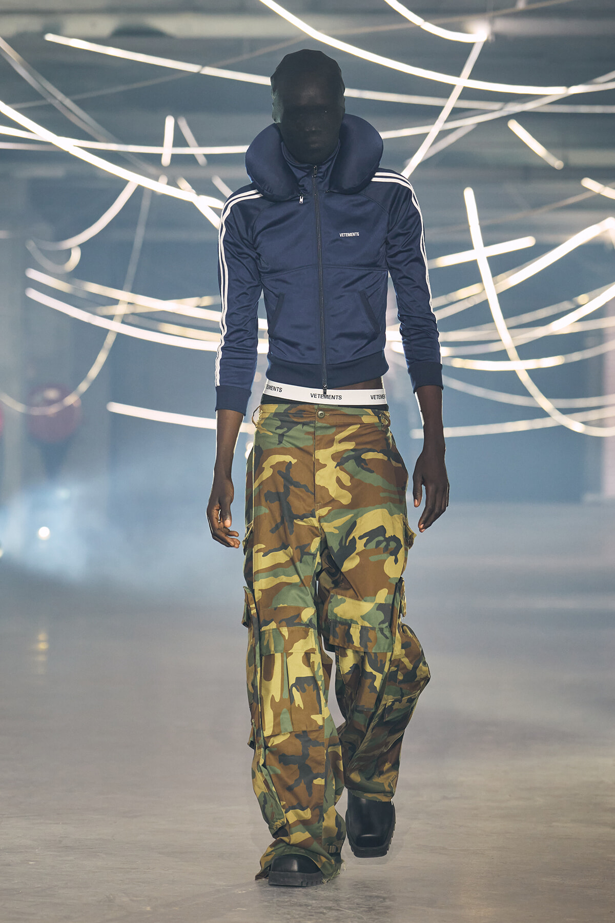 Male model walking the runway in a navy zip-up jacket and camouflage cargo pants, with neon light tubes overhead.
