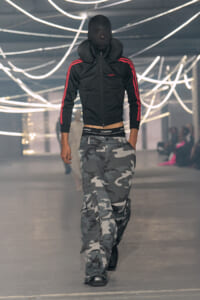 Male models walks a fashion runway wearing a black zip jacket with red stripes and gray camouflage cargo pants under bright, looping lights.