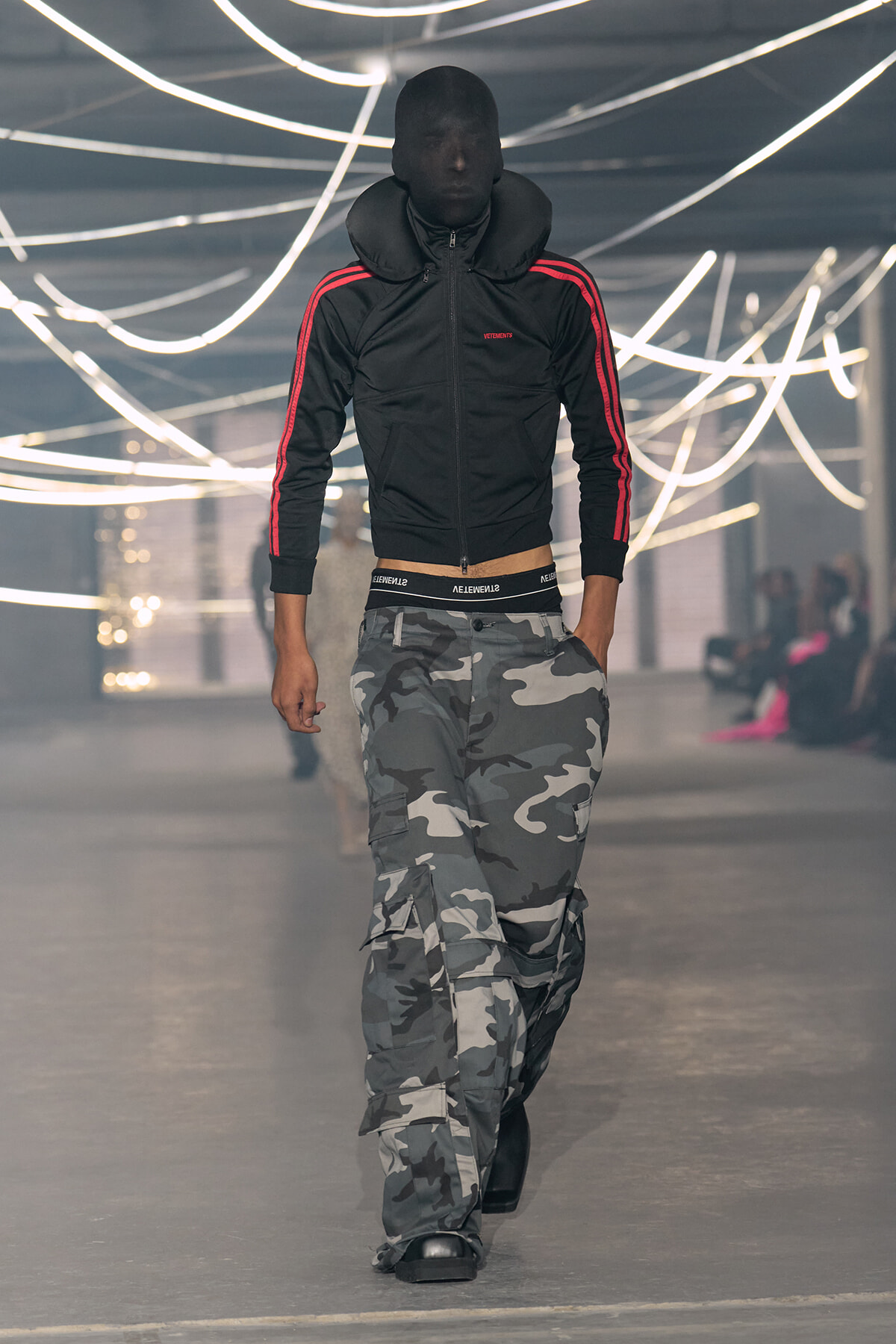Male models walks a fashion runway wearing a black zip jacket with red stripes and gray camouflage cargo pants under bright, looping lights.