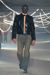 Male model walking a fashion runway in a glossy black jacket over a beige shirt with faded jeans and black shoes, neon light decor in the background.