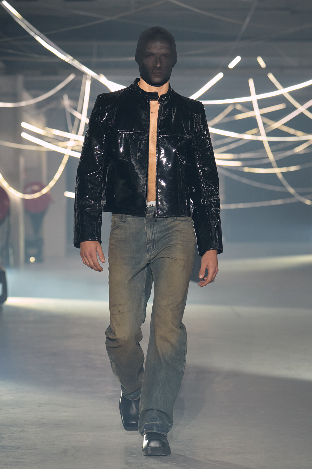 Male model walking a fashion runway in a glossy black jacket over a beige shirt with faded jeans and black shoes, neon light decor in the background.