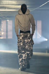 Person wearing a light gray zip hoodie and camouflage cargo pants walks toward the camera in a hazy, industrial space.