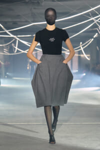 Model walking a fashion runway in a black T-shirt with small white text and a voluminous gray midi skirt, with black tights and open-toed shoes; neon lights in the background.