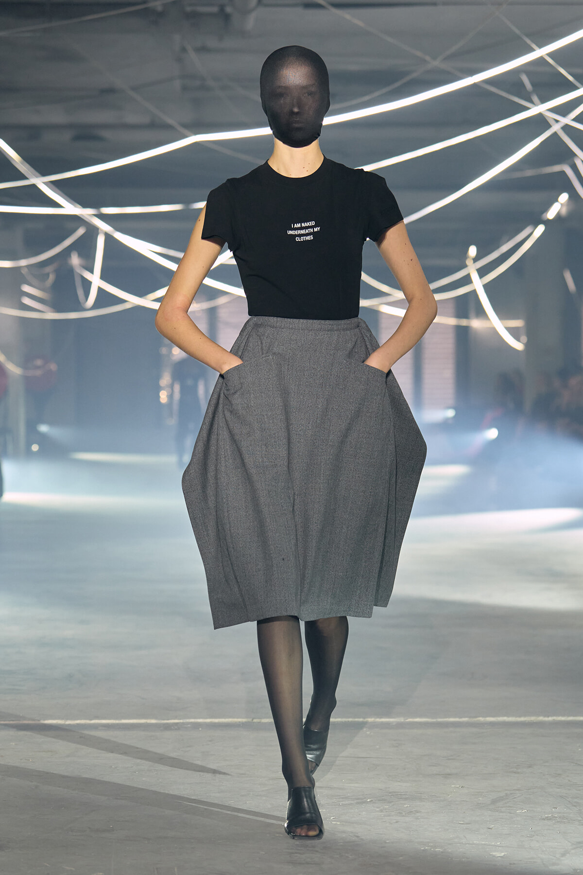 Model walking a fashion runway in a black T-shirt with small white text and a voluminous gray midi skirt, with black tights and open-toed shoes; neon lights in the background.
