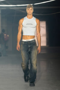 Male runway model wearing a light gray cropped tank top with text, loose jeans, and black boots, his head covered with a neutral mask during a fashion show