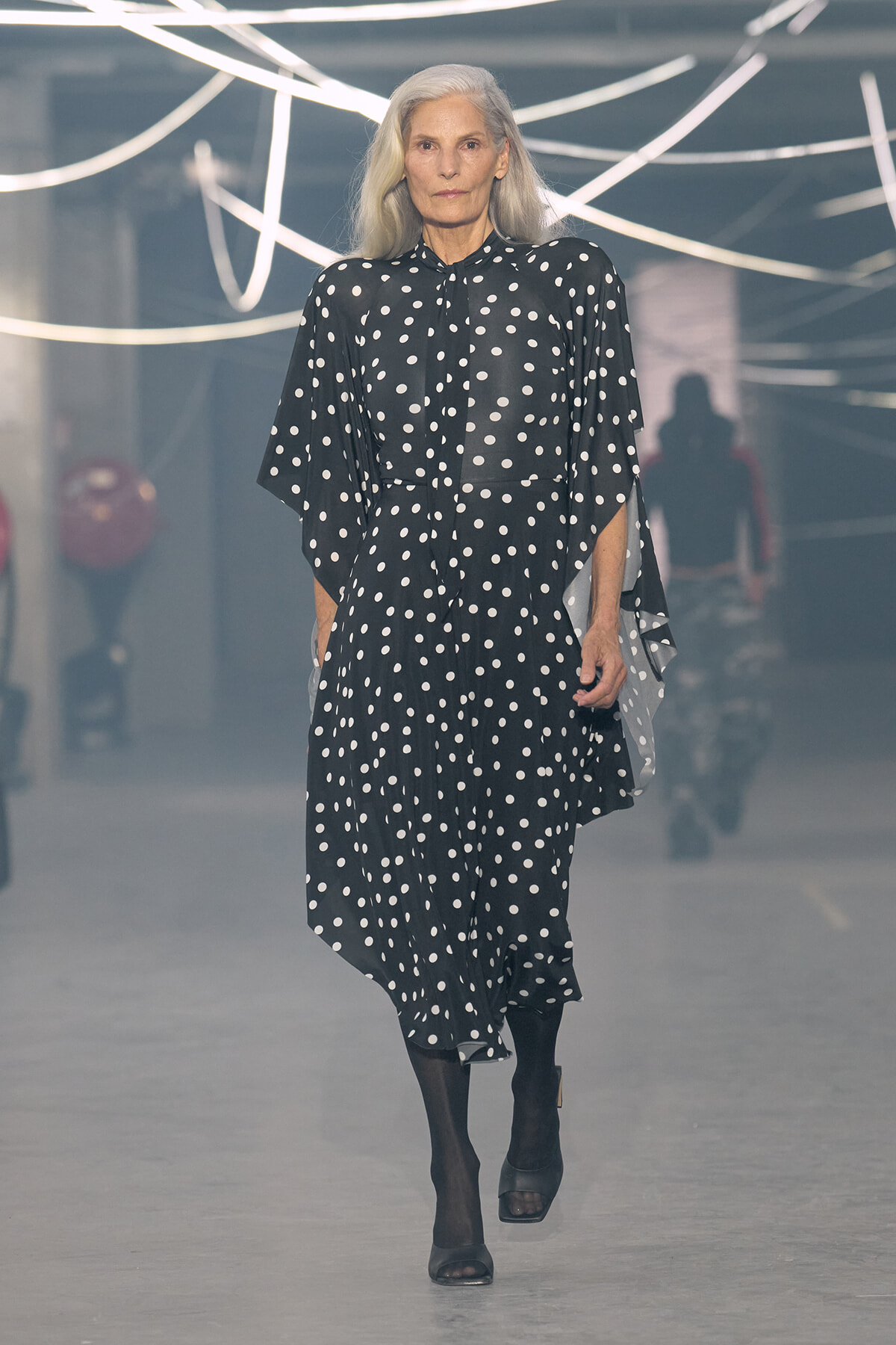 Model walking a fashion runway in a black polka-dot dress with wide sleeves and black footwear.