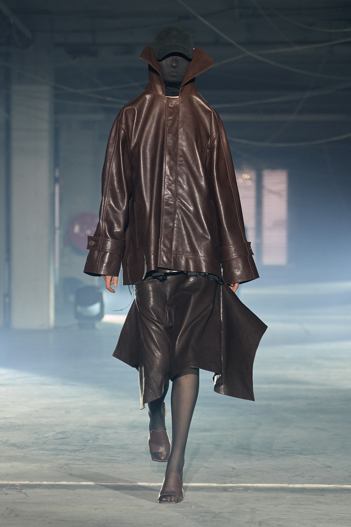 Model walking a runway in an oversized brown leather coat with a high collar and matching skirt, black tights, and open-toe sandals, dim venue lighting.