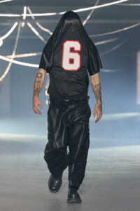 Person walking away on a runway, wearing a black jersey with the number 6 and hood over their head.