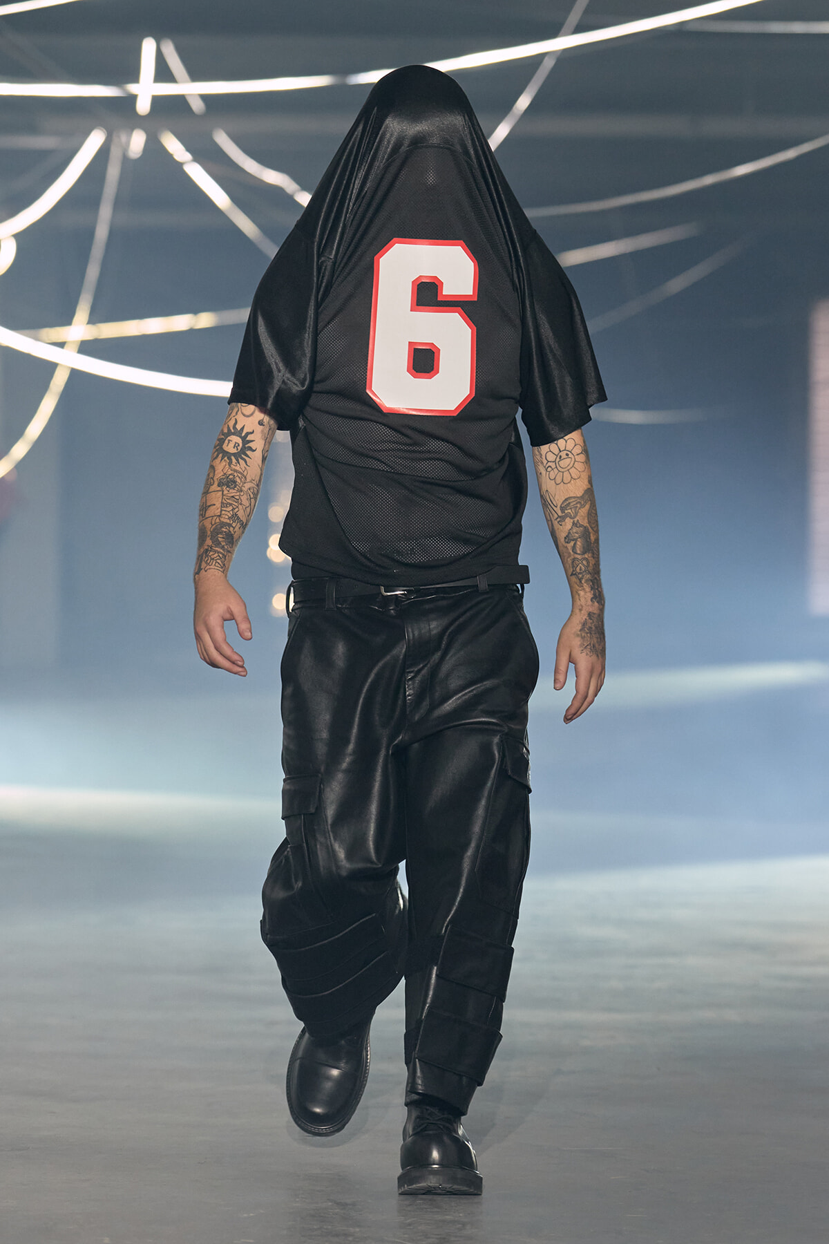 Person walking away on a runway, wearing a black jersey with the number 6 and hood over their head.