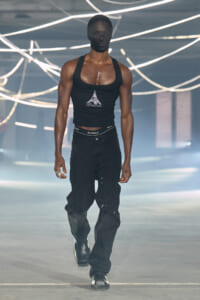 Male model wearing a black balaclava, sleeveless top with Eiffel Tower graphic, and baggy jeans walks a runway under neon lights.