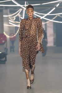 Model walking a fashion runway in a brown polka-dot dress with sheer puff sleeves and matching heels.
