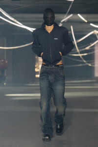 Male model walking the runway in a dark zip-up hoodie, baggy jeans, and black boots, with his face covered by a black balaclava.