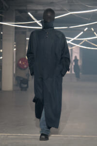 Tall figure wearing a long black oversized coat with a high collar, walking down a runway in an industrial space with glowing neon tubes in the background.