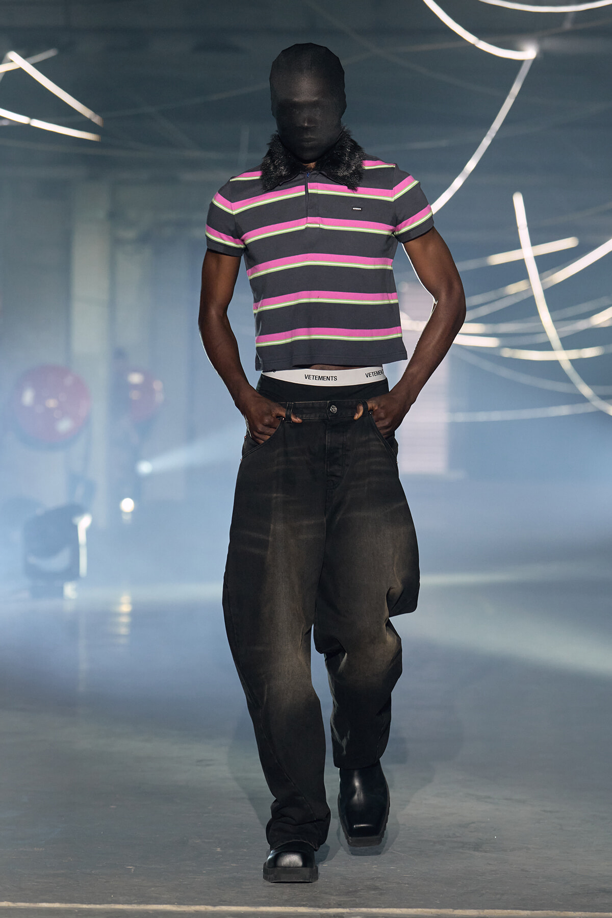 Male model walking a fashion runway in a striped polo shirt and oversized dark jeans, with a black balaclava and neon light backdrop.