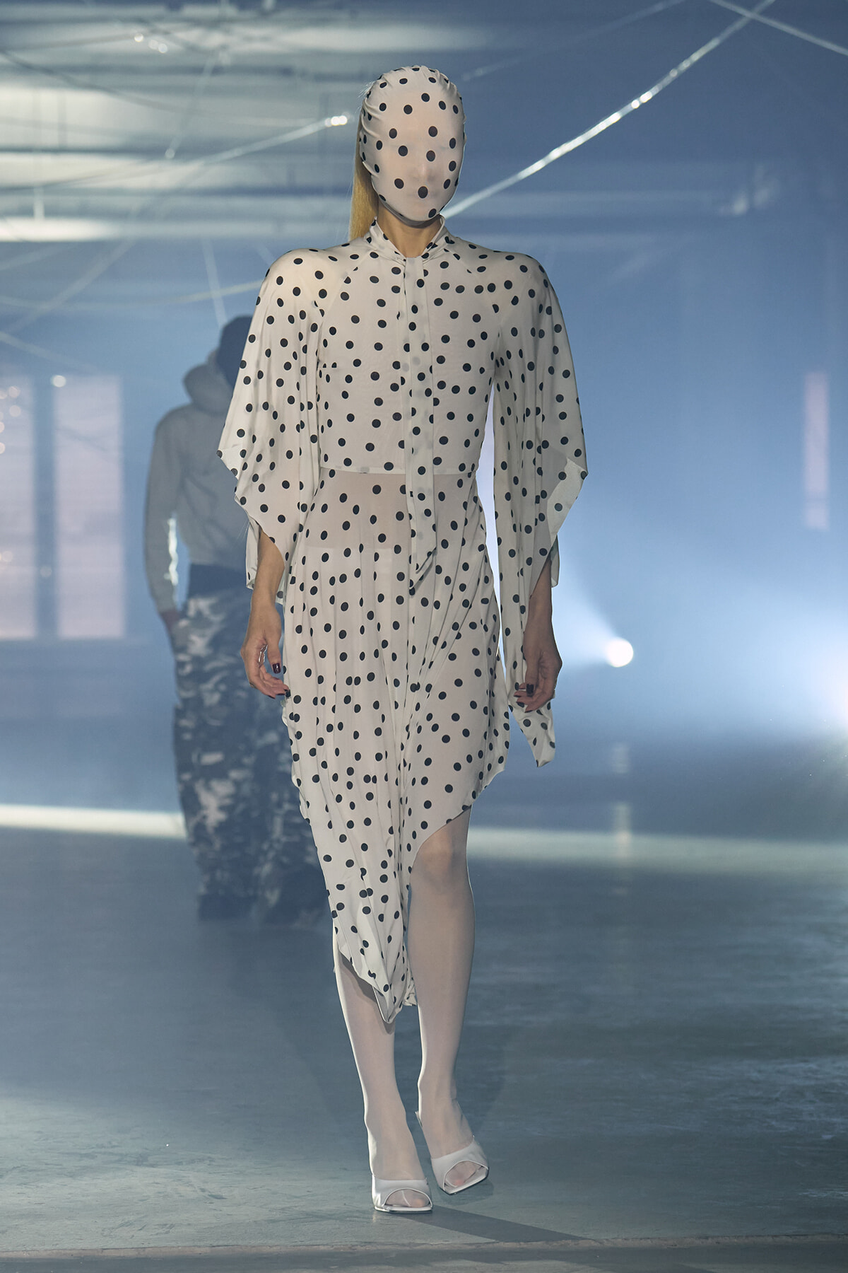 Model in a white polka-dot dress and matching headscarf walks the runway amid foggy blue lighting at a fashion show
