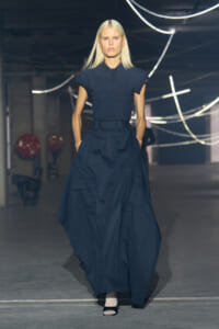 Model walking the runway in a dark navy polo tucked into a voluminous cargo skirt, hands in pockets, industrial backdrop with neon tubes.