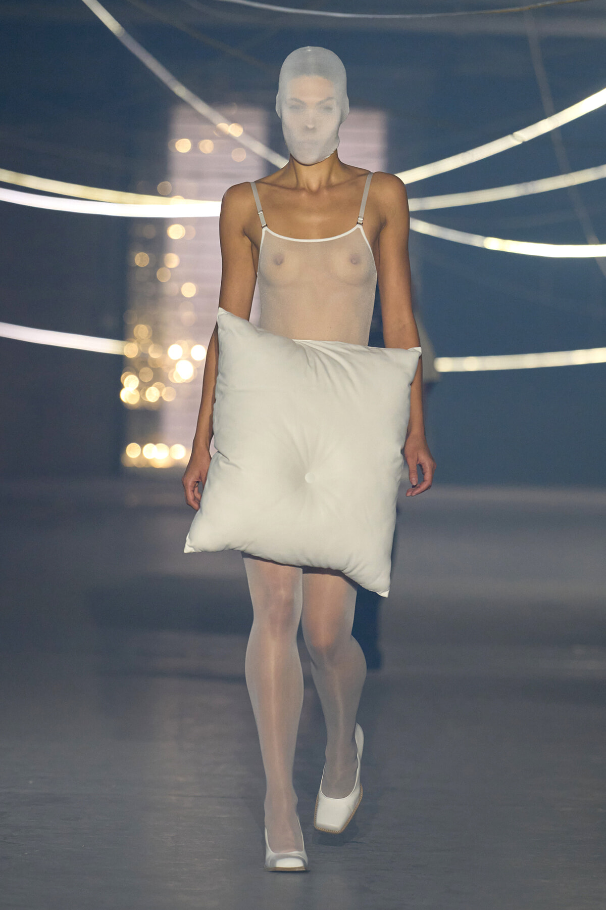 Model walking a runway in a sheer white slip dress and veil, carrying a large white pillow.