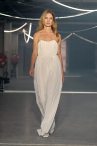 Model walks a runway in a strapless white, flowing gown with a cinched waist under bright neon lights.
