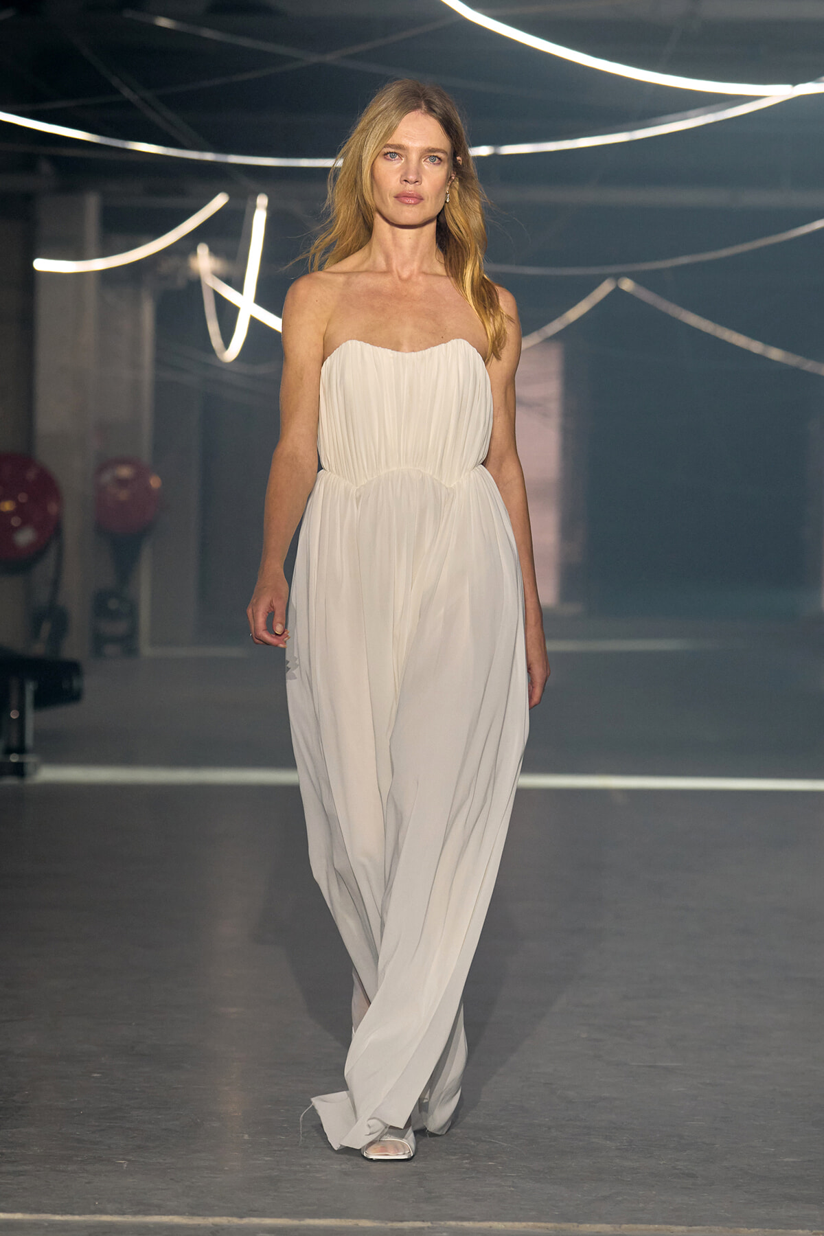 Model walks a runway in a strapless white, flowing gown with a cinched waist under bright neon lights.
