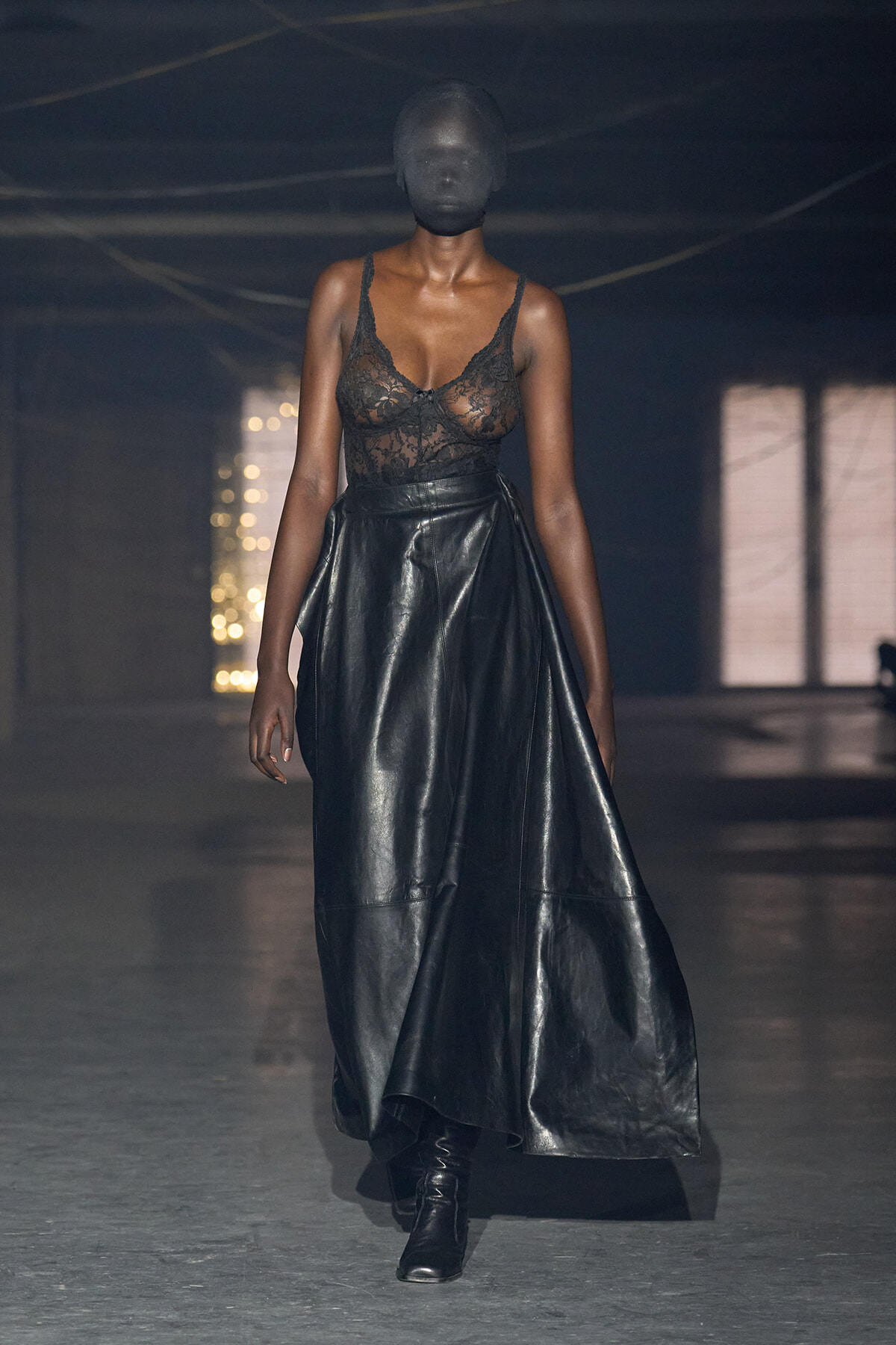 Model walking a dimly lit runway in a sheer black lace bodice, full black leather skirt, and black boots; face fully covered by a dark hooded mask.