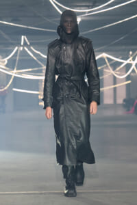 Model walks the runway in a black leather trench coat with belt, hood up, in a neon-lit industrial setting.