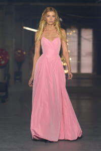 Female model walking a floor-length pink halter-neck evening gown on a fashion runway, with a soft-lit backstage backdrop behind her.