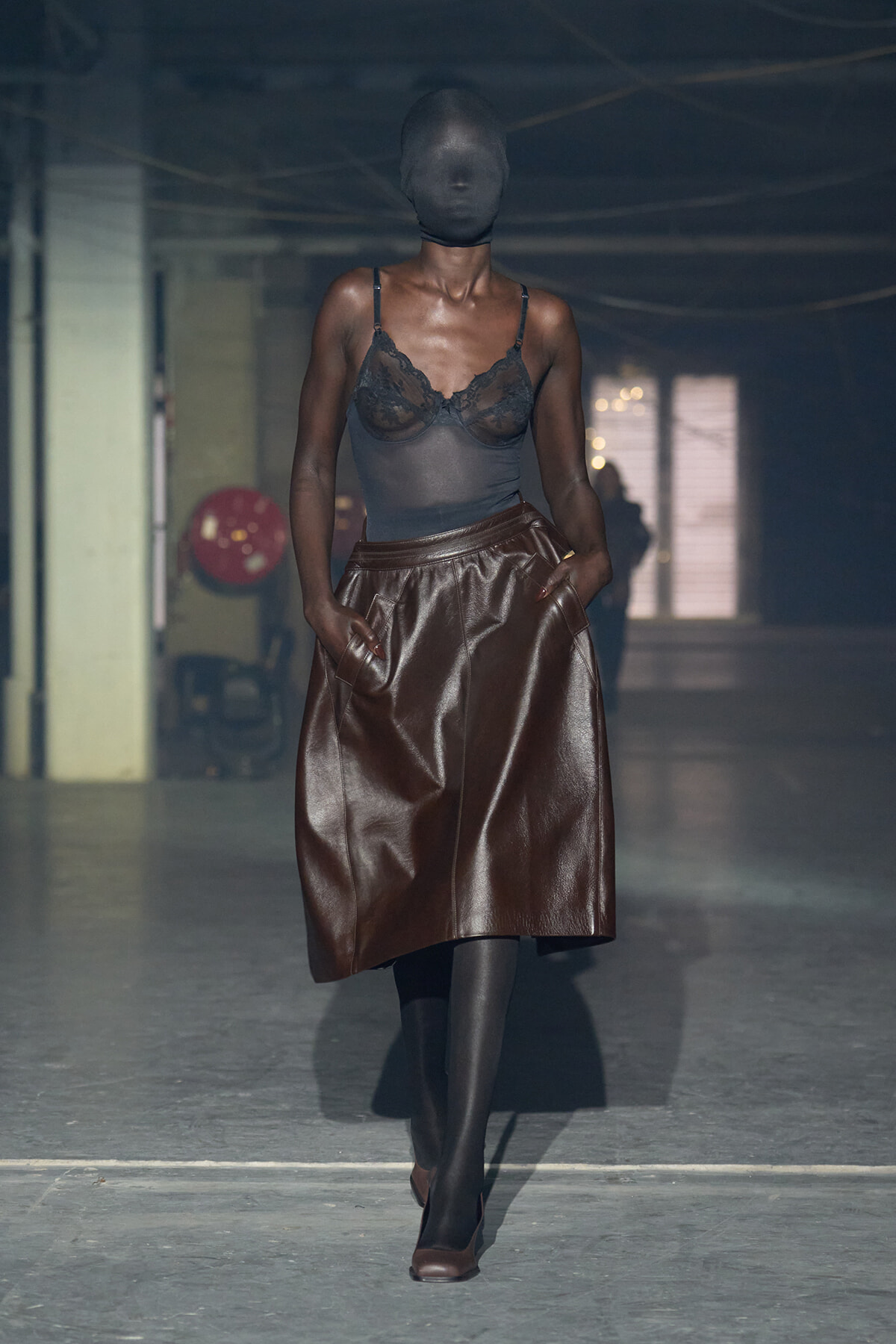 Runway model wearing a sheer black lace top tucked into a high-waisted brown leather midi skirt, with a full-face black hood and black tights.