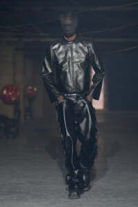 Model walking a dimly lit runway in a full black leather ensemble: jacket and pants, with a black balaclava-style face covering.