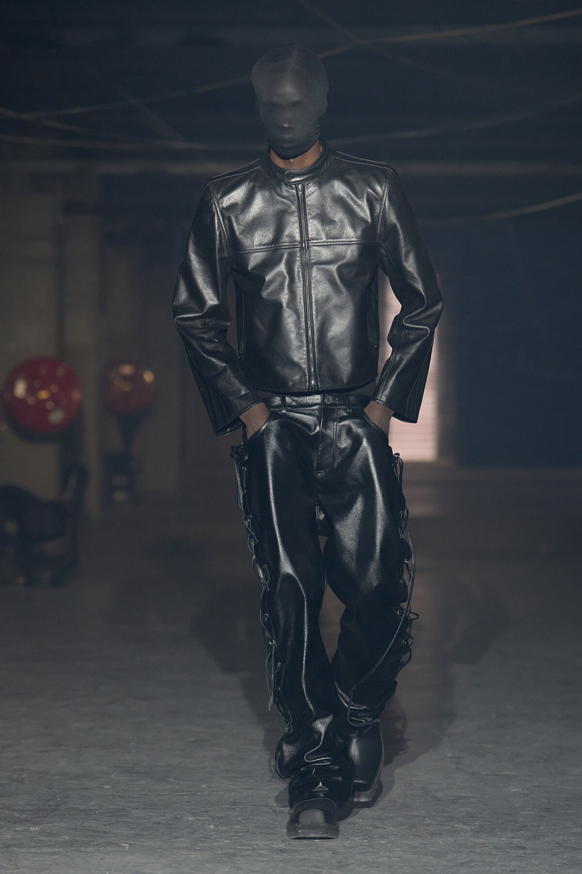 Model walking a dimly lit runway in a full black leather ensemble: jacket and pants, with a black balaclava-style face covering.