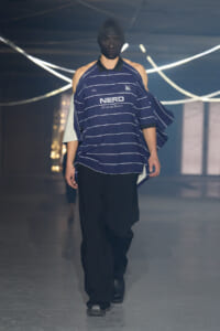 Model walking the runway in a navy striped oversized polo with a black face mask, paired with black wide-leg pants and black boots.
