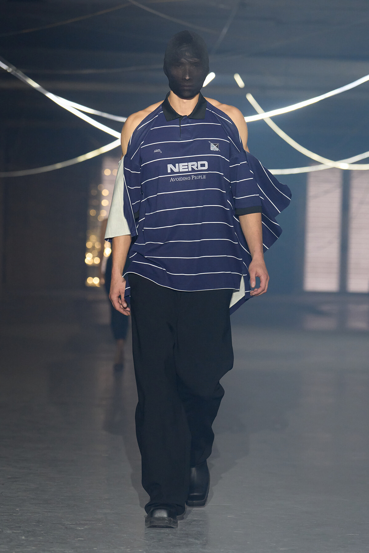 Model walking the runway in a navy striped oversized polo with a black face mask, paired with black wide-leg pants and black boots.