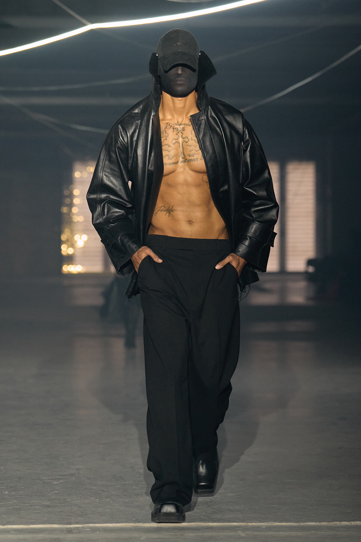 Male model on a runway wearing an open black leather jacket, black cap, and black pants, exposing a tattooed chest.