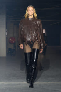 Model walks a runway wearing a brown leather fringe jacket and black thigh-high boots.
