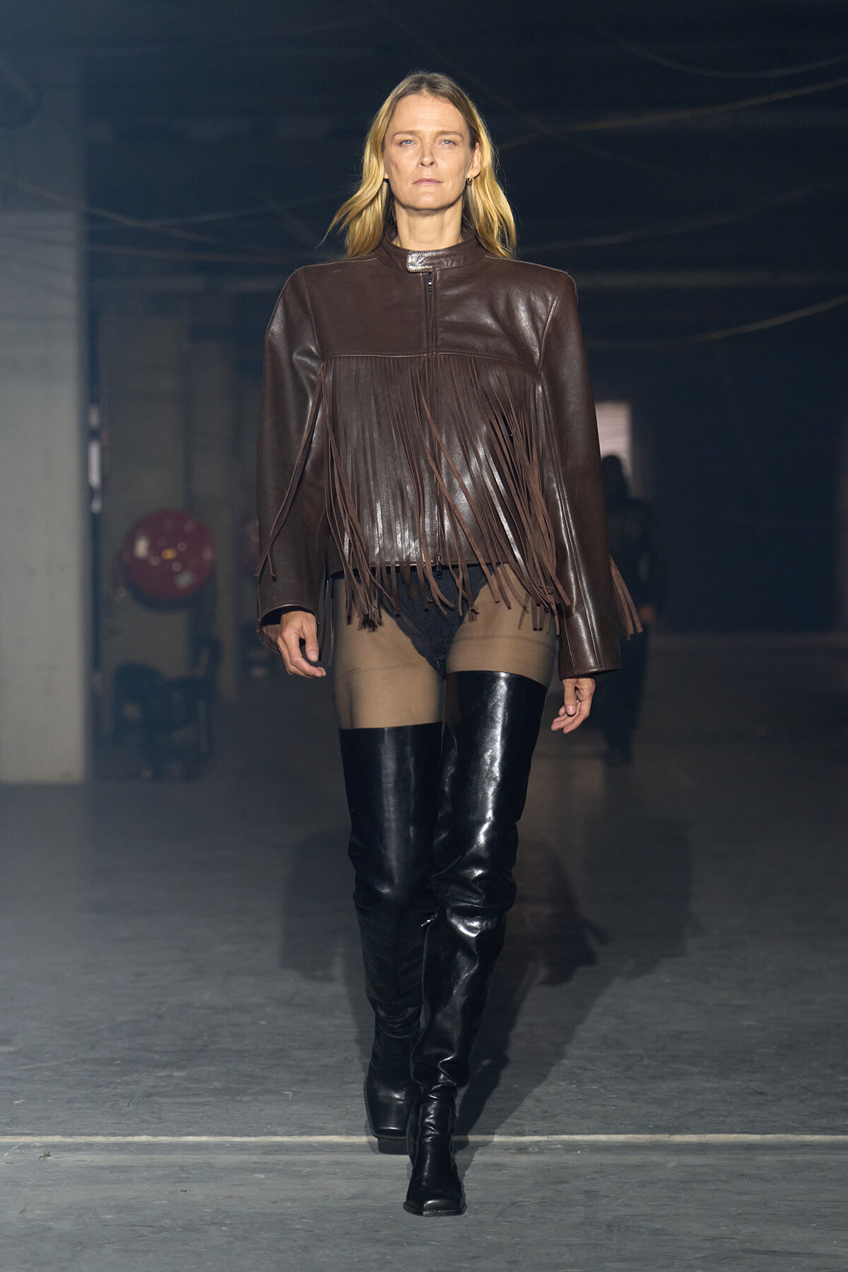Model walks a runway wearing a brown leather fringe jacket and black thigh-high boots.