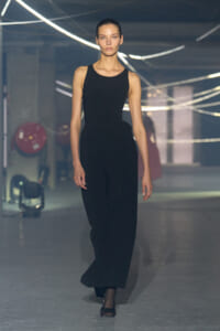 Model walking on a runway in a sleeveless black jumpsuit, reaching camera level as she strides forward.