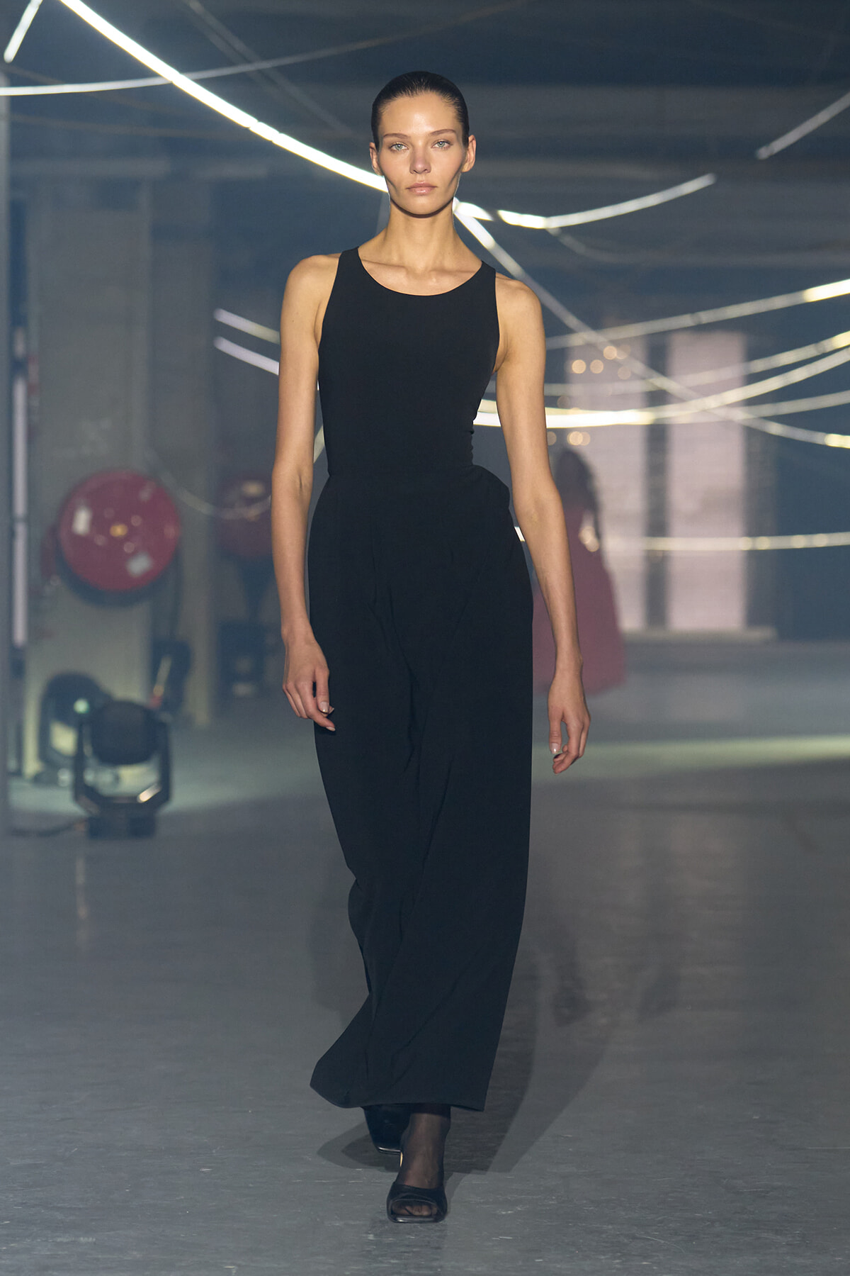 Model walking on a runway in a sleeveless black jumpsuit, reaching camera level as she strides forward.
