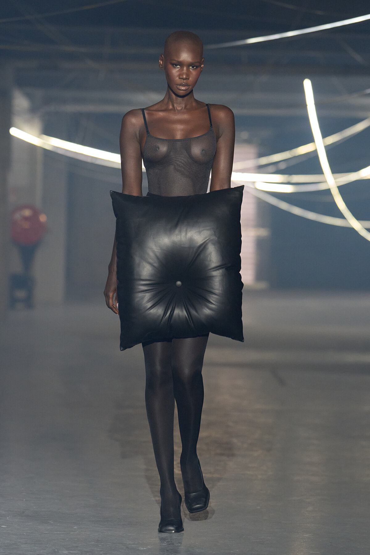 Model walks the runway wearing a sheer black top and opaque tights while holding a large black square cushion.
