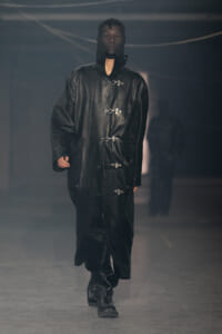 Model walking the runway in a long black leather hooded coat with metal closures on the front.