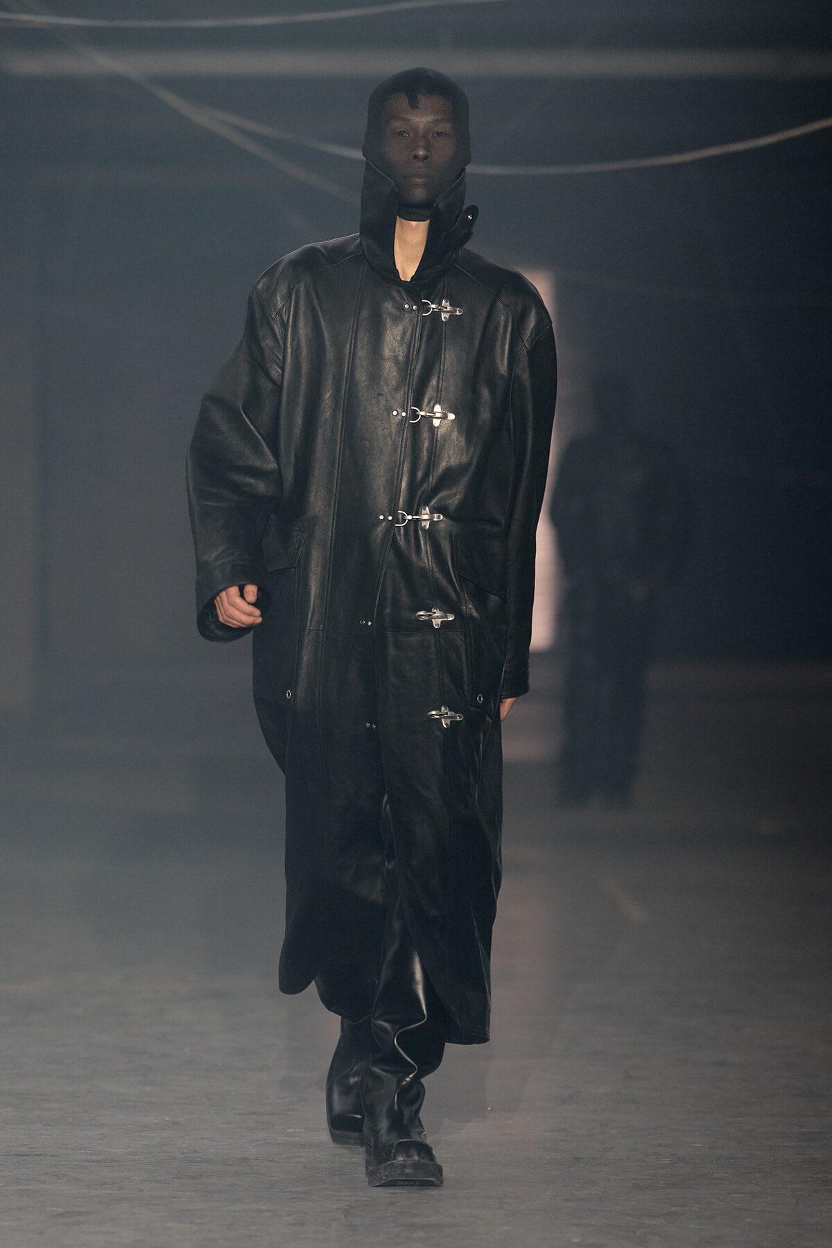 Model walking the runway in a long black leather hooded coat with metal closures on the front.