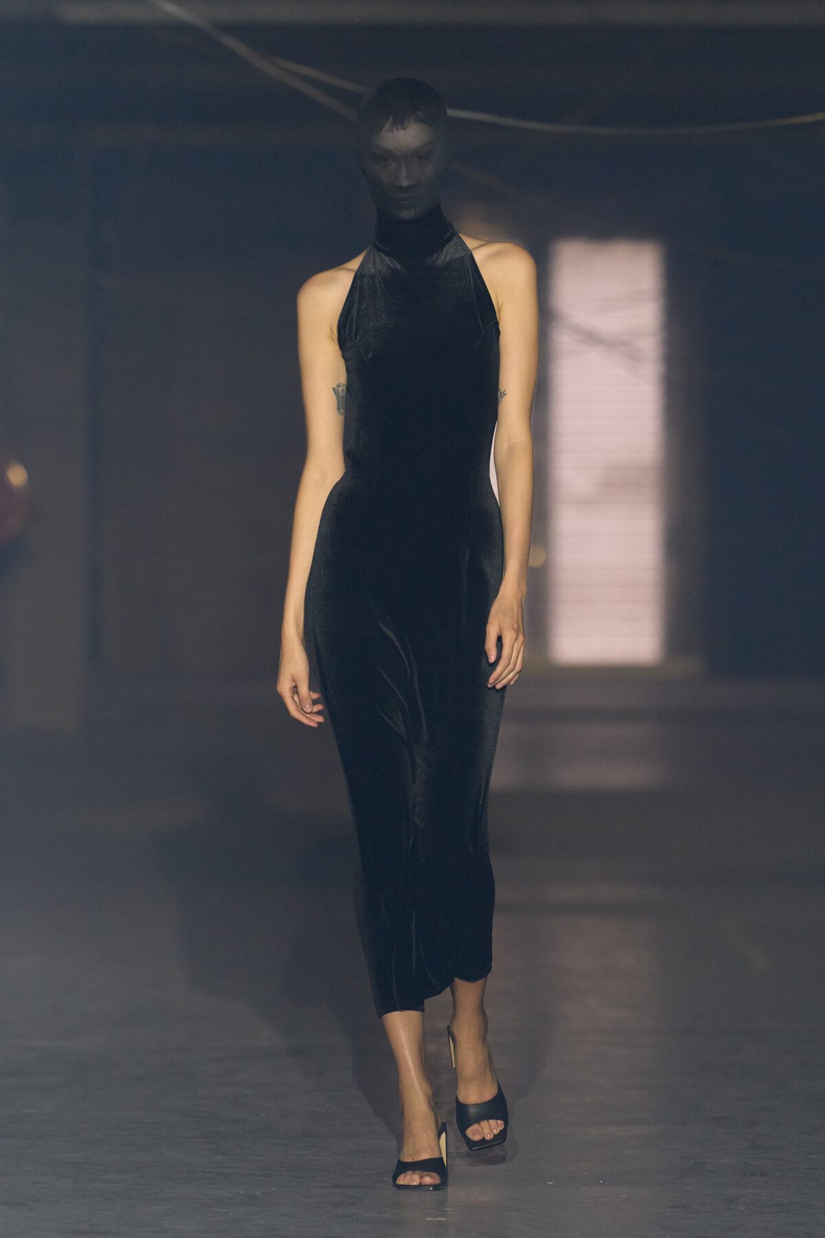 Model walking the runway in a black velvet halter-neck dress; head and face obscured by a black mask or hood.