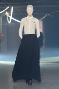 Model in a white button-down shirt and a floor-length black maxi skirt walks a fashion show runway, face obscured by a head covering.