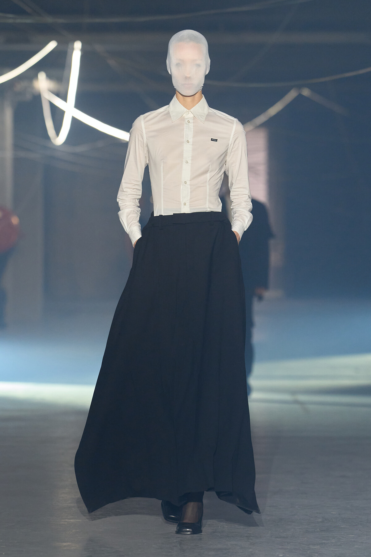 Model in a white button-down shirt and a floor-length black maxi skirt walks a fashion show runway, face obscured by a head covering.