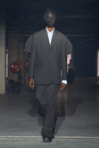 Male model walks a runway in an oversized gray suit with a white shirt, dim industrial backdrop behind him.
