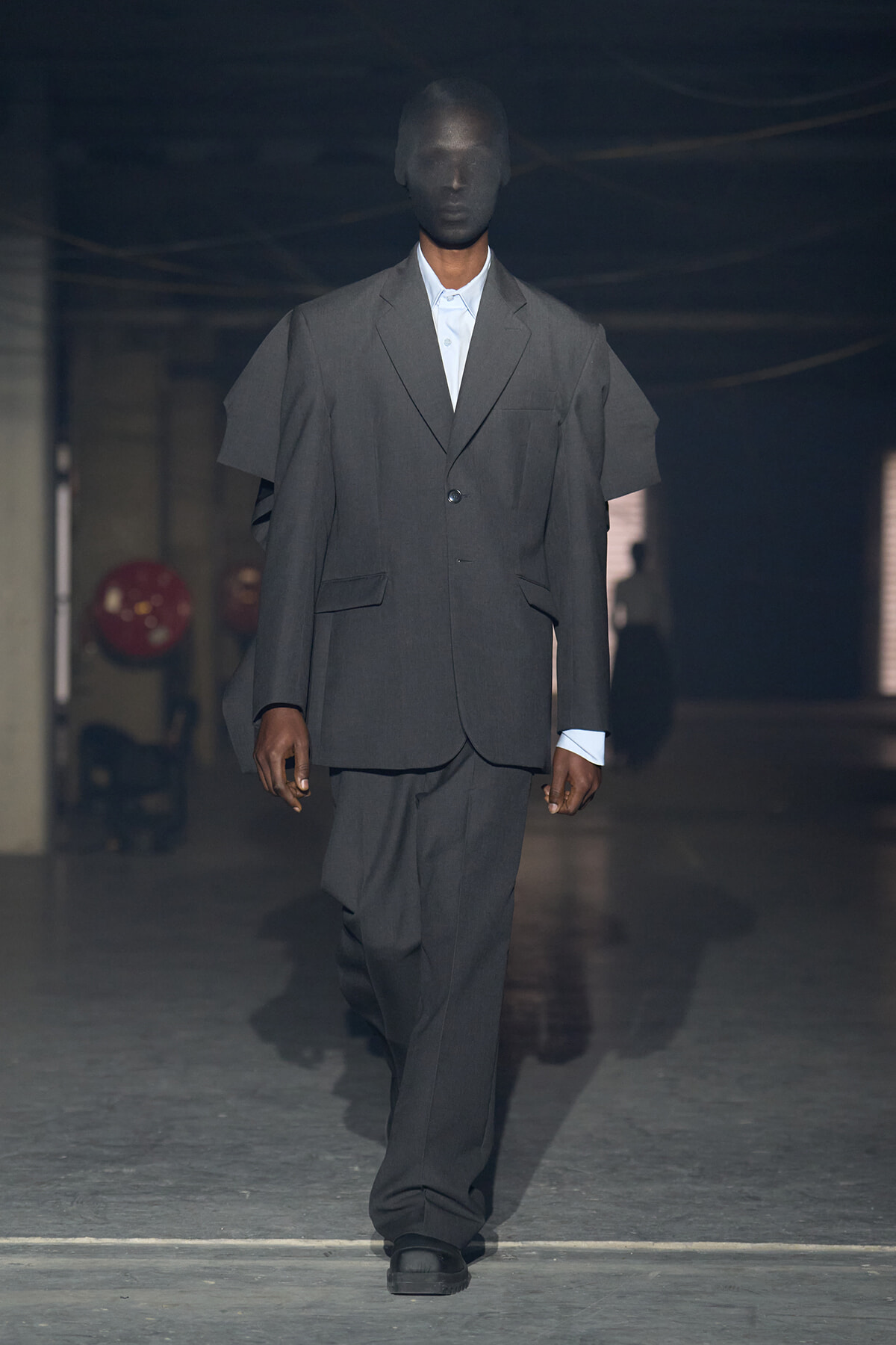 Male model walks a runway in an oversized gray suit with a white shirt, dim industrial backdrop behind him.