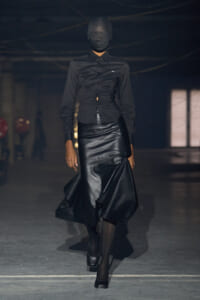 Runway model in an all-black outfit: button-down blouse with a fitted, sculpted leather skirt; head wrapped in black fabric.