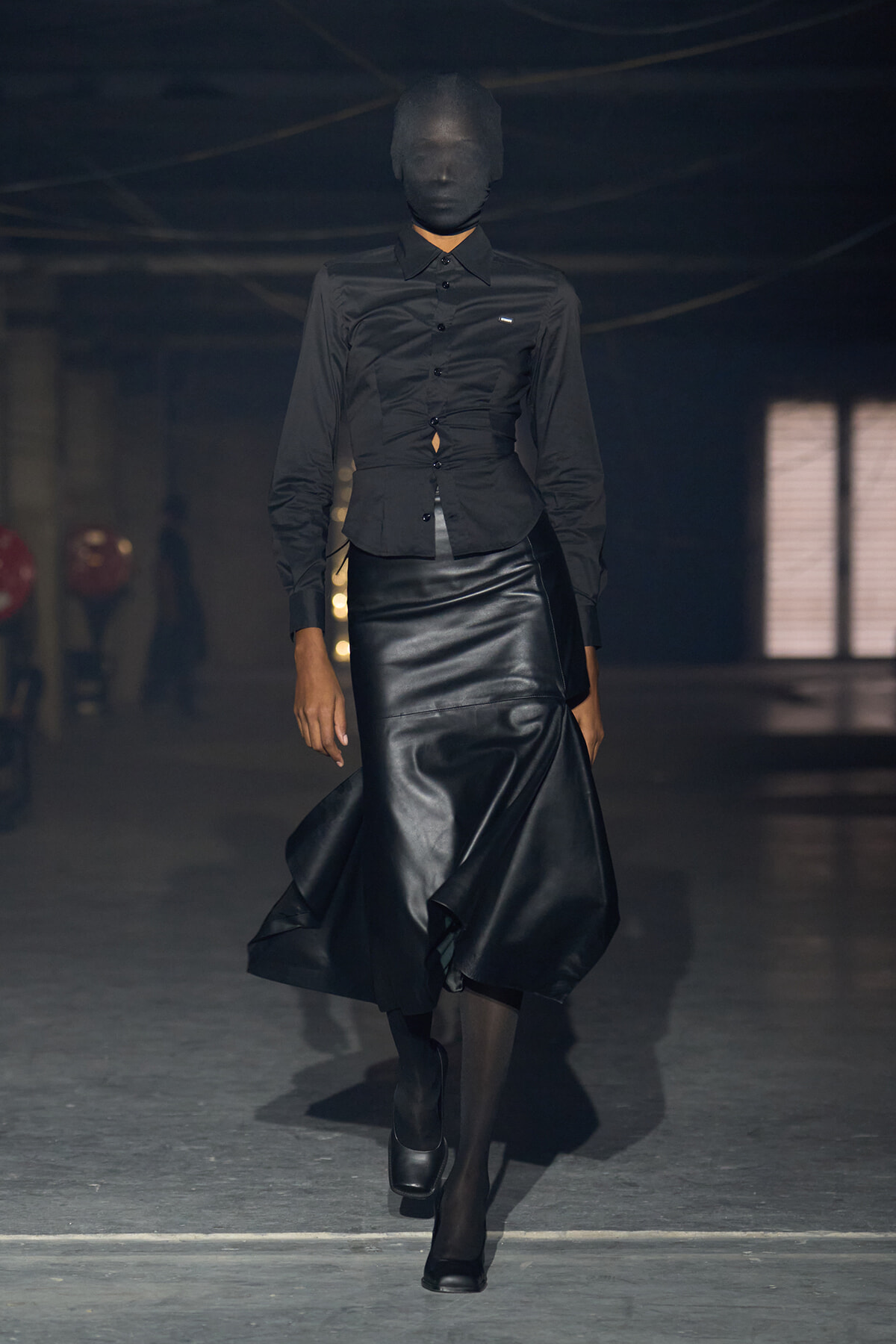 Runway model in an all-black outfit: button-down blouse with a fitted, sculpted leather skirt; head wrapped in black fabric.
